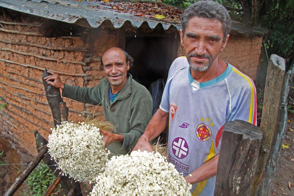 Two middle age men stand in front of a house holding large bouquets of flowers.