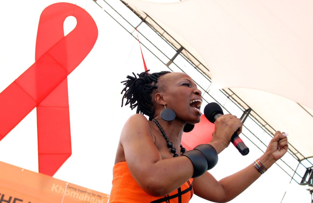 A Black woman sings in front of the HIV red ribbon symbol.