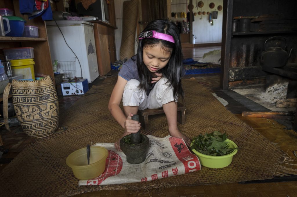 A young girl pounds a mortar into a pestle, inside a house.