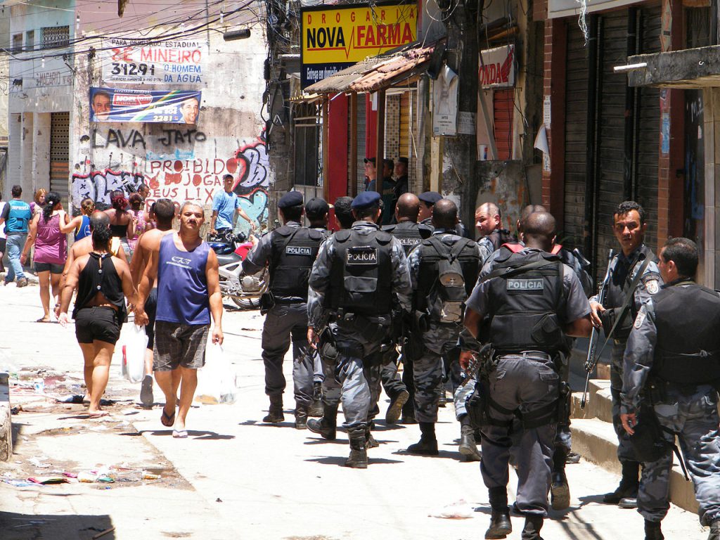 The photograph features several police officers uniformed in black walking past casually dressed people on a public road lined with shops.