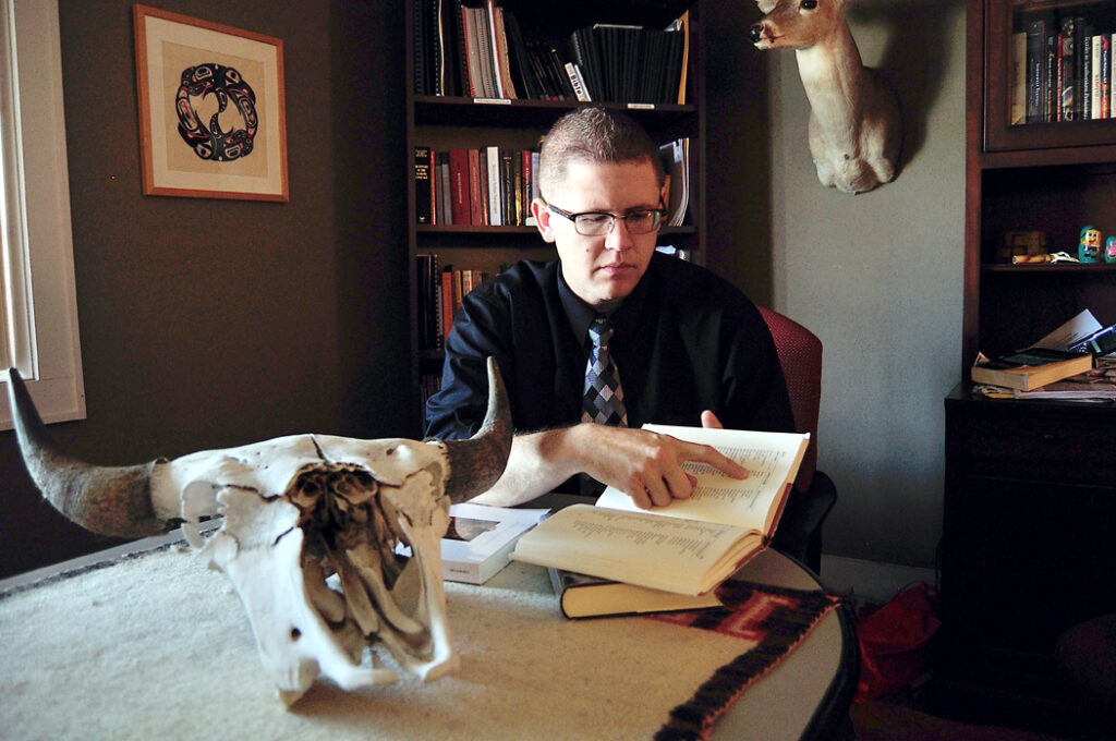 A man sits at a desk and points to words in a book. Behind him are bookshelves, and a skull rests on the desk in front of him.