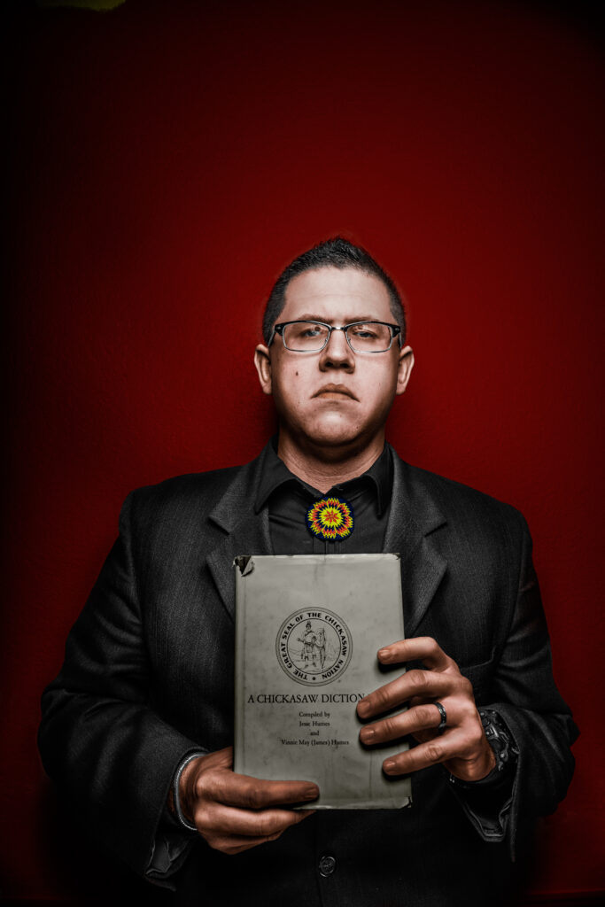 A man wearing a jacket holds a book and stands against a red background.