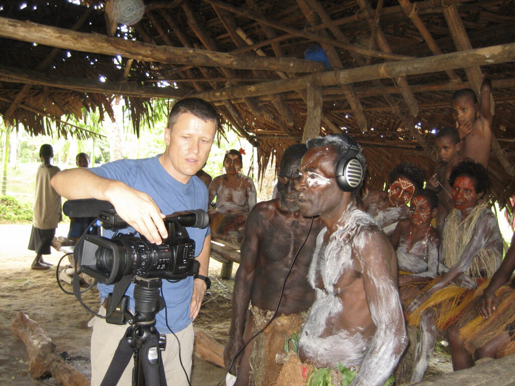 A man on the left shows two men on the right how to work a video recorder. The men are gathered in front of a group of seated people.
