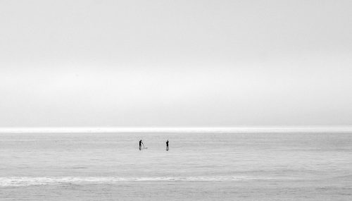 Two people on paddle boards paddle off into a gray ocean covered by a gray sky.