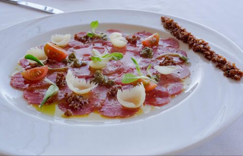 This dish is an Italian-inspired baby alpaca carpaccio with pasankalla, herb-infused oil, Parmesan scoops, and sliced cherry tomatoes.