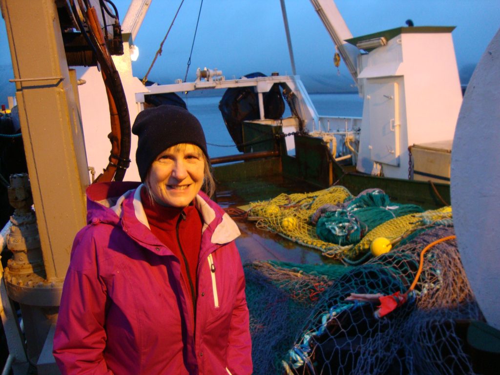A woman in red coat and black hat stands on a fishing boat.