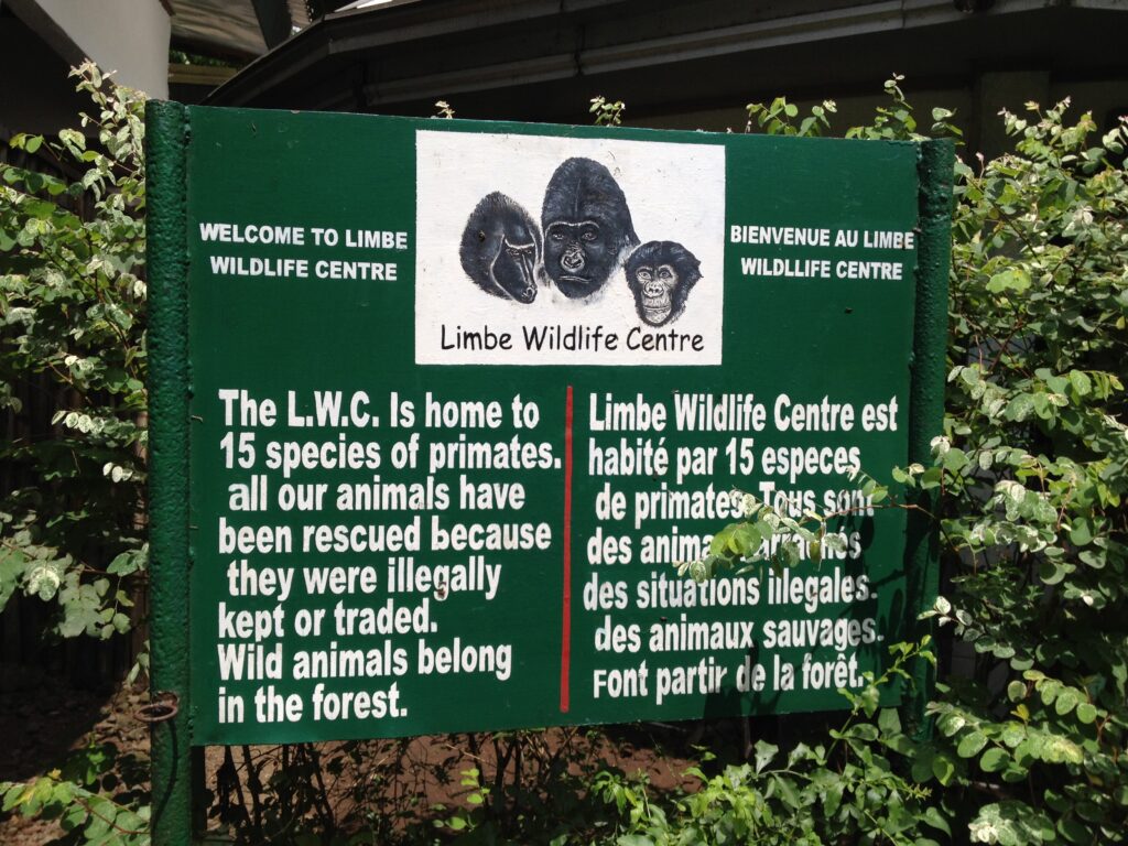 A mostly green sign surrounded by shrubbery has an illustration of the faces of three primates in a white box above text that reads, “Limbe Wildlife Centre.” Below this box, white text reads, “Welcome to Limbe Wildlife Centre. The L.W.C. is home to 15 species of primates. All our animals have been rescued because they were illegally kept or traded. Wild animals belong in the forest.” To the right of this text and a red dividing line is its French language translation.