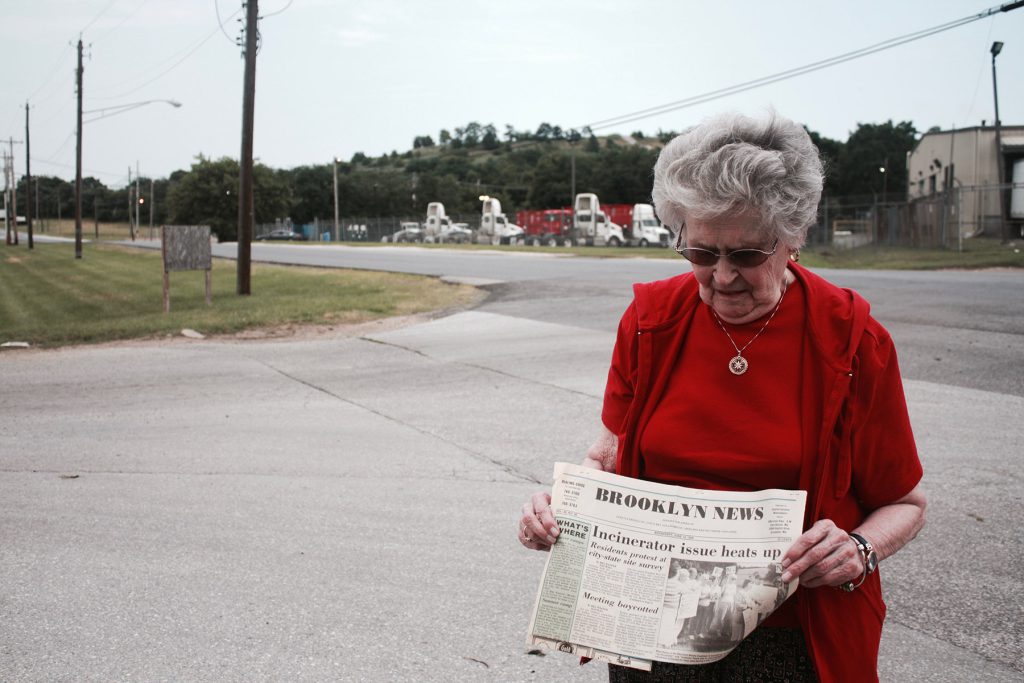 Minnie stands outside the nation’s largest medical waste incinerator and shows the author an article from the 1980s about protests against the plant that her husband helped to organize.
