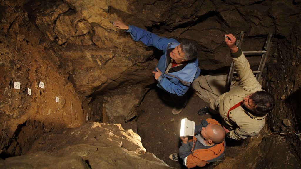 Three researchers stand in Denisova Cave in Russia, home to the newly classified Denisovan skull fragments.
