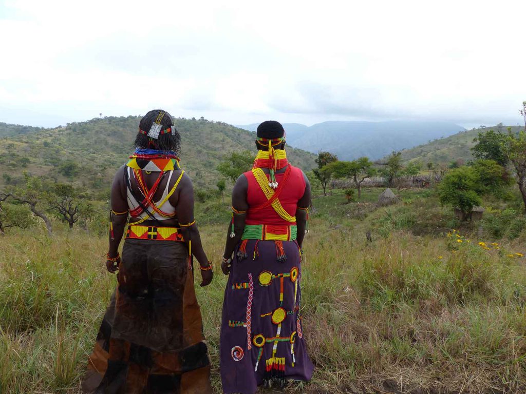 Two Ik women in traditional dress walk through a field in Uganda.