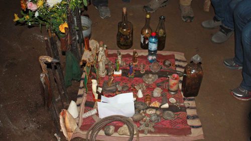 A photo shows a table with figurines, stones, candles, shells, papers, and other ritual objects scattered across a colorful woven cloth.