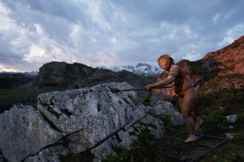 On a mountainous landscape with sunlight shining in from the top right corner, a naked person with short brown hair holds a spear horizontally with both hands and points it ahead of them.