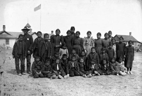Some of the Lakota children from the previous photo appear in this image after they entered the Lower Cut Meat Creek Indian Day School. At the beginning of each school year, students’ familial clothes were replaced with uniforms, the boys’ hair was cut, and pupils were issued Anglicized names. Note the hand of policeman Lee Wood firmly on the shoulder of student Eddie Foot.