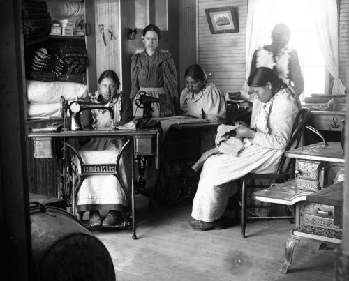 native american boarding schools - Native American girls were trained to be productive, subservient homemakers in the mold of Victorian norms. In this photo, Bratley’s wife, Della, teaches dressmaking to Lakota students Gracie Good-Bird, Emma Elk Looks Back, Nellie Foot, and Bessie Elk Looks Back (left to right). Native American schools were often staffed with husband-and-wife teachers who acted as surrogate parents, enticing children away from their biological families and toward a model of Anglo-American domesticity.