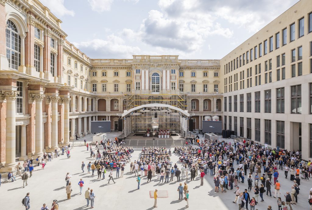 The photograph features many people in a sunny, open plaza with buildings towering above on three sides.