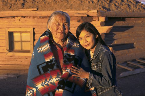 A photo shows a Navajo elder wrapped in a blanket, standing next to a Navajo girl dressed in a jean jacket.