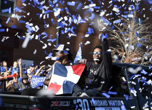 Two men ride in the back of a truck while blue and white confetti flies through the air and a crowd below cheers.