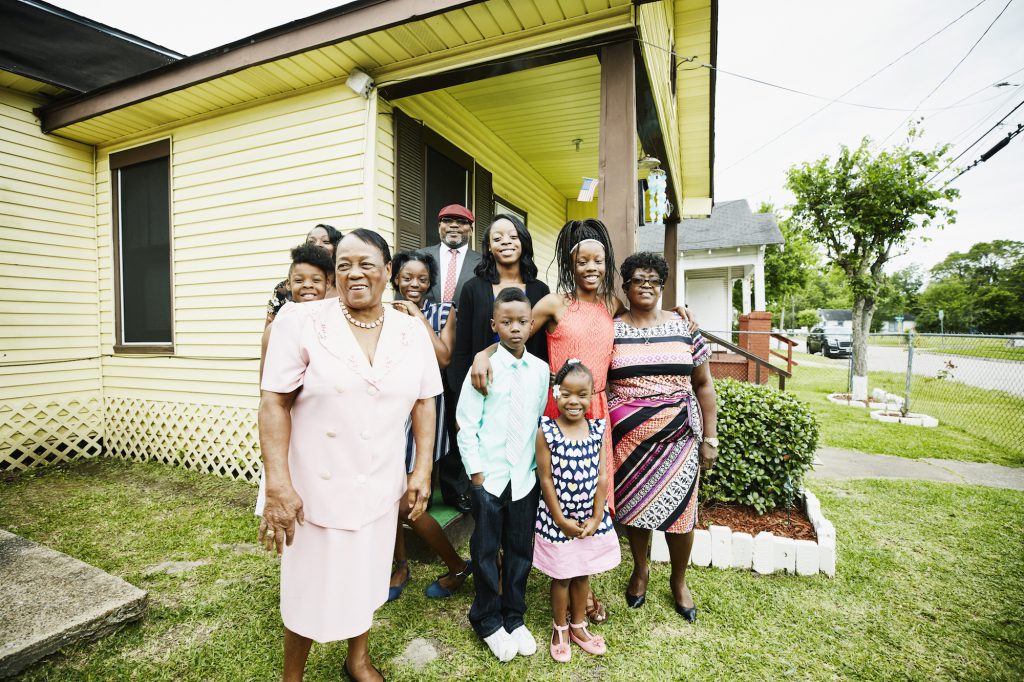 A group of people of various ages stands in front of a house, smiling at the camera.