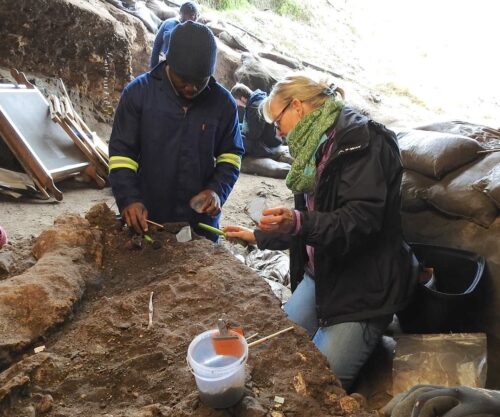 A black man in a blue jump suit and a white woman in a coat and scarf kneel in the dirt with tools excavating