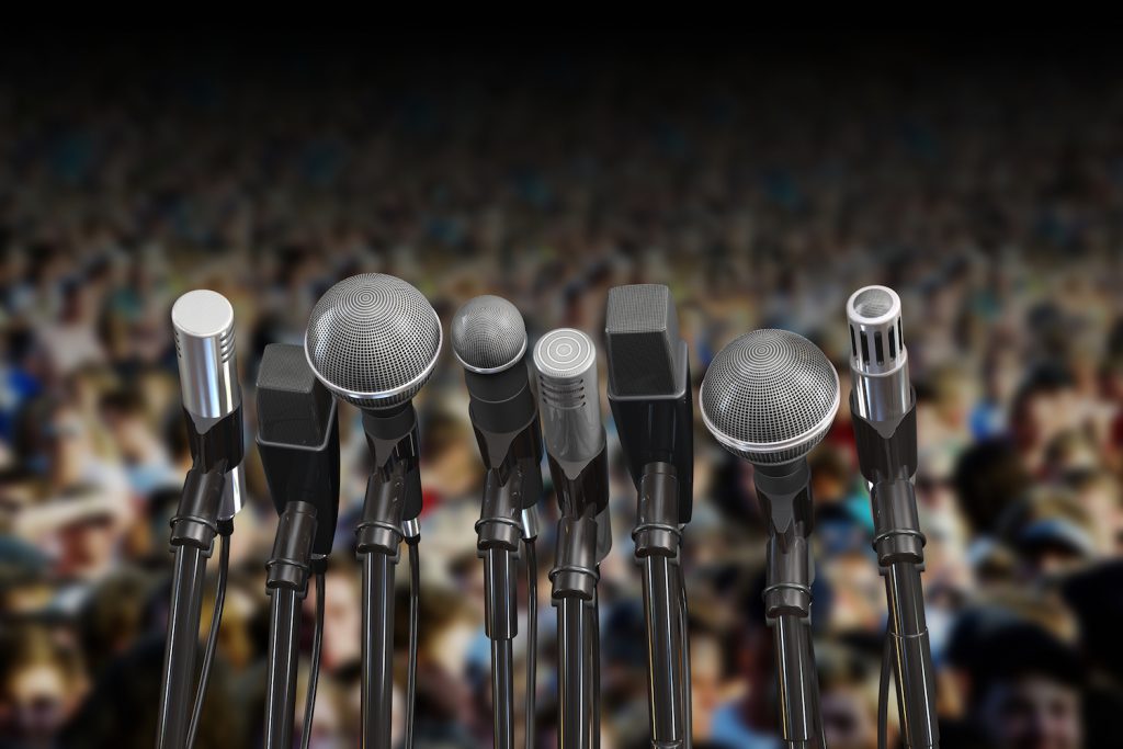 A photo shows microphones lined up together in front of a blurred out crowd of people.