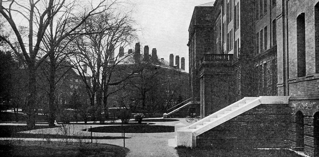 Stairs lead up to a large brick building on the left, with other brick buildings in the background.