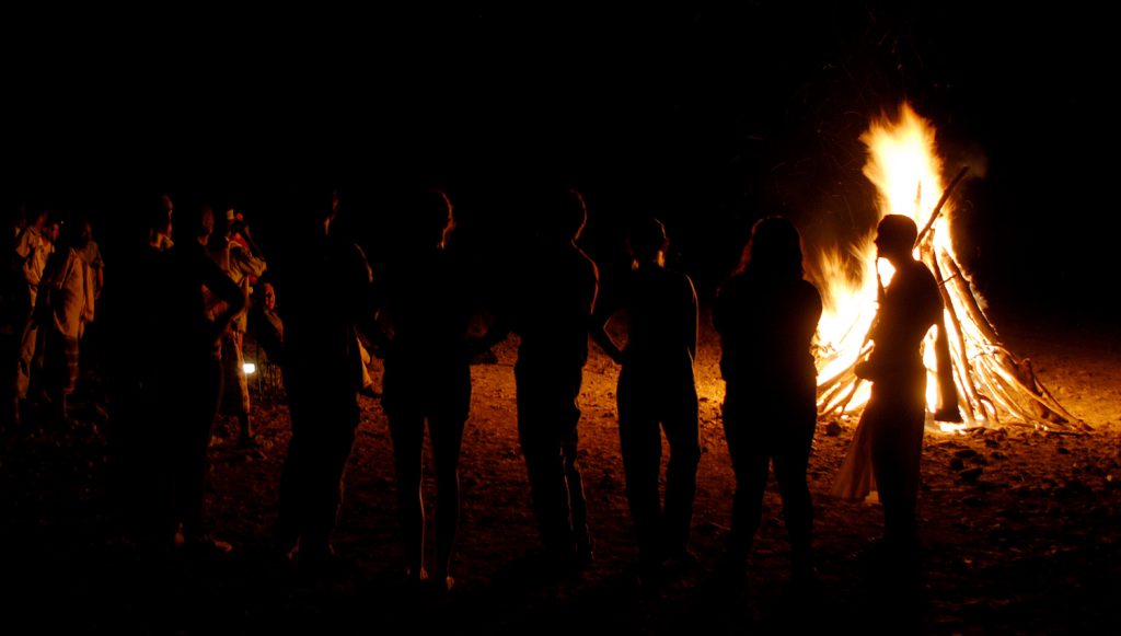 A group of people stand with their backs to the camera around a bonfire at night