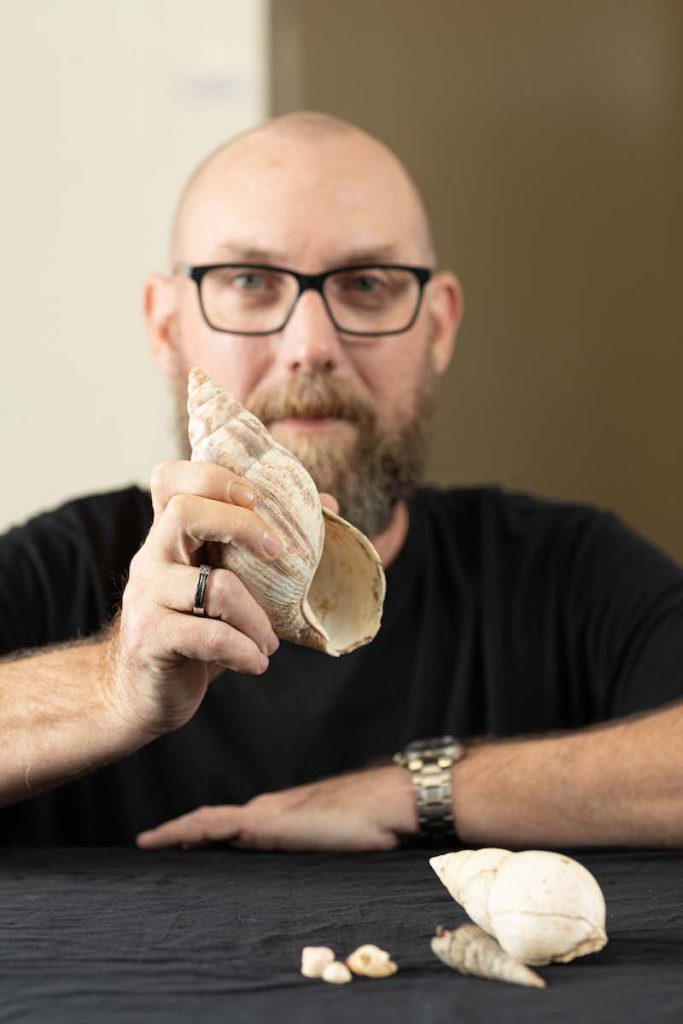 A man with a shaved head, black glasses, and a beard wearing a black T-shirt sits at a table with different sized, light colored shells on it. He holds up a giant snail shell in his right hand.