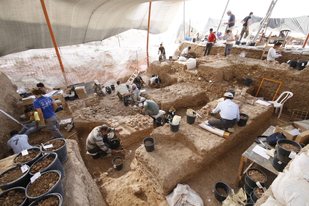 Several people dig and sift through brown dirt, surrounded by black buckets and tools.