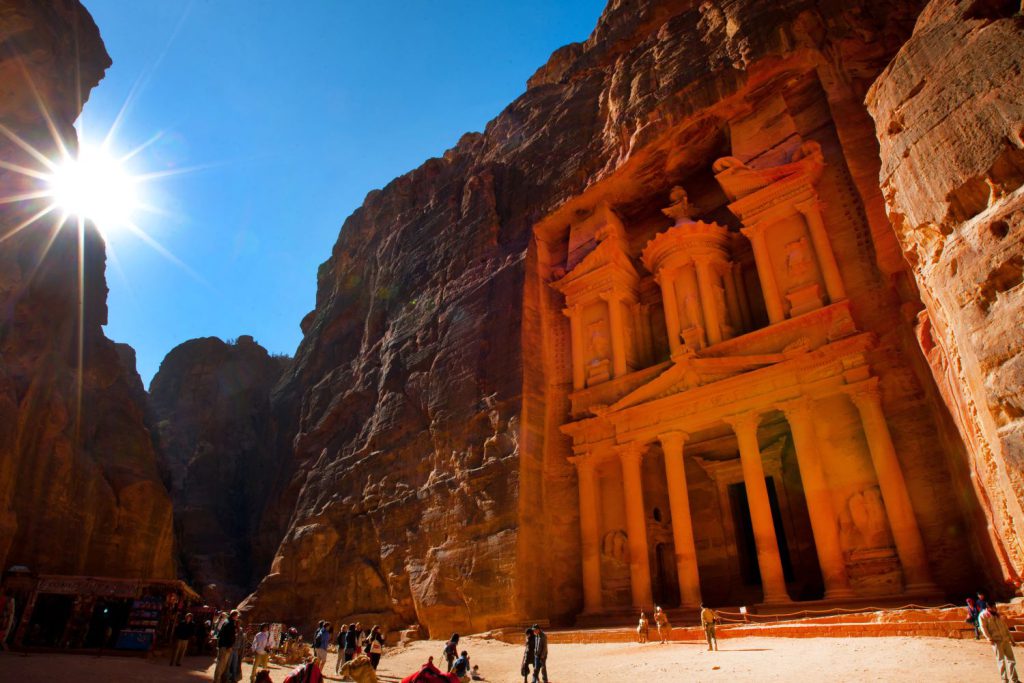 In a valley, an archaeological site is carved into a mountainside, at the base of which tourists gather. A bright sun and blue sky are in the background.