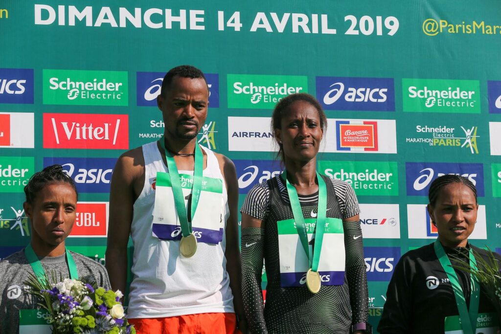 Four athletes stand on a winner’s podium with medals around their necks. The people on the left and right are both holding flowers.