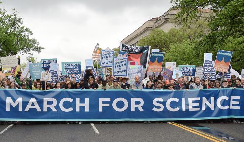 A large group of people walk down an avenue holding signs behind those who carry a long blue-and-white banner that reads “March for Science.”