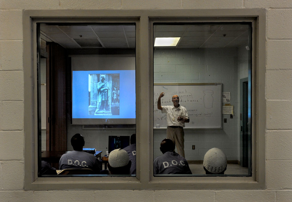 Through two windows, a teacher in a white shirt stands in a classroom in front of a whiteboard and beside a screen with a slide on it. Four students wearing blue shirts and two with white caps on face the teacher with their backs to the windows.