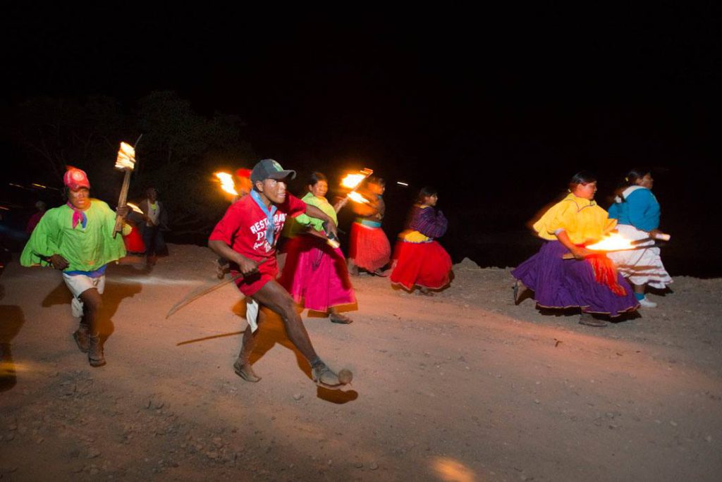 Men and women in bright, colorful clothing run across sands at night, carrying torches.