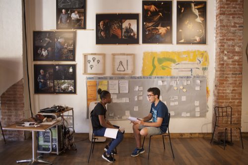 Two young people sit facing each other with paper on their laps. Behind them are large photos and a large grey storyboard.