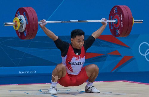 A male Olympic weightlifter snatches a weighted barbell.