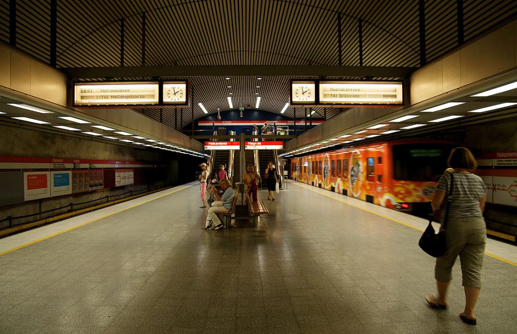 People sit and stand near a bench on an underground subway platform flanked by two sets of train tracks. A train moves through the station on the right.