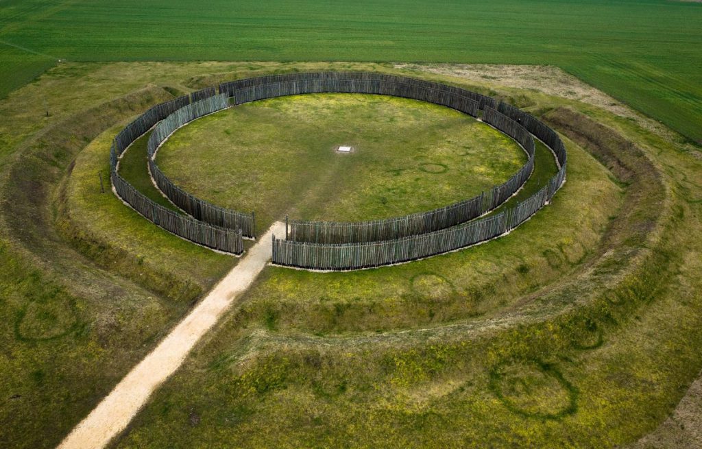In a green field of grass, a large mound of earth rises. On the mound, two circles of wooden fences with openings are encircled by a ditch.
