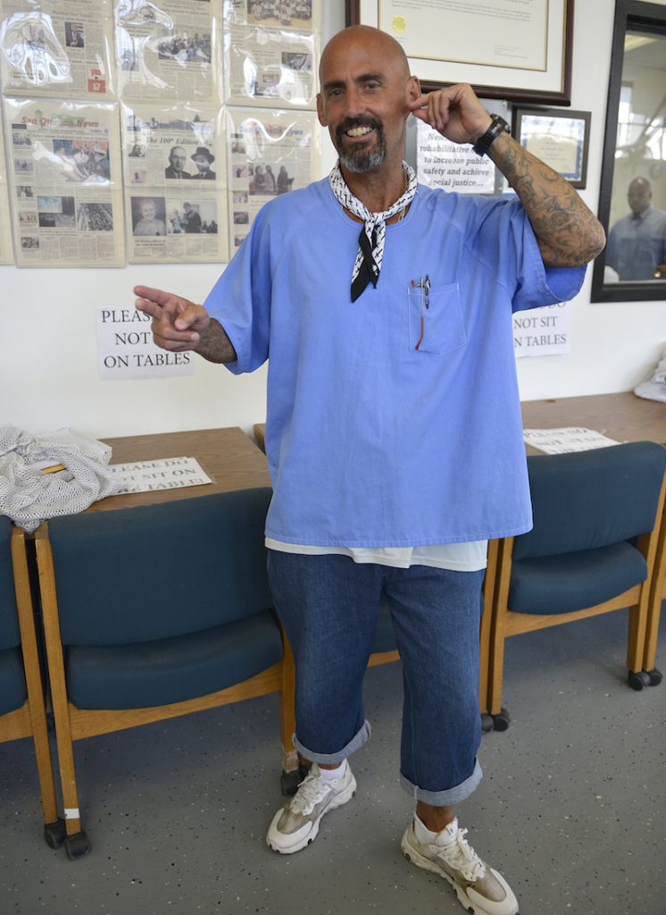 A man with a bald head wearing a large blue shirt, blue jeans, and a bandana tied around his neck pulls his left ear with one hand and extends the other, holding out two fingers in the handshape of the letter "h" in American Sign Language.