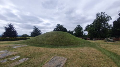 A grass-covered mound, used by the Anglo-Saxons, sits surrounded by trees and hedges, with flat, concrete slabs on the ground in front of it.