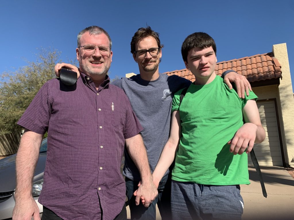 A father and son hold hands, while a man stands behind them with his arms around both of them.