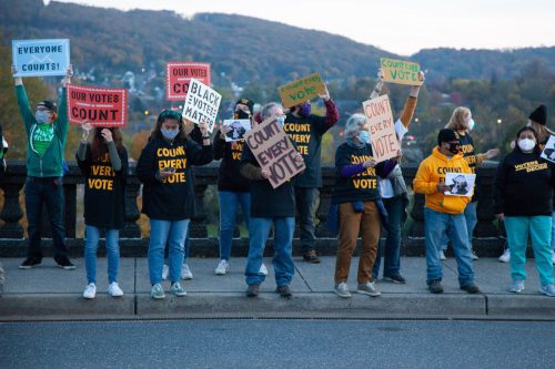 A group of people hold up signs on the side of a road. Their shirts and signs say “count every vote.”