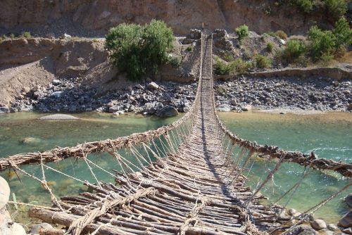 A woven rope bridge extends out over a river to the other side.