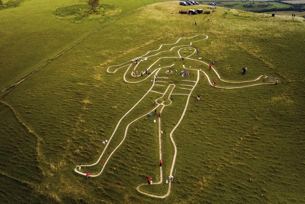 An aerial image shows a large chalk drawing of a nude man holding a large stick carved into a hill. A group of people are spread out and cleaning different parts of the chalk drawing.