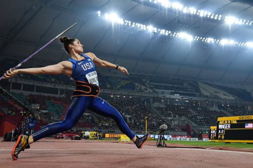 An athlete in mid-stride dressed in blue tights and a tank top prepares to throw the javelin in a large, well-lit sports arena.