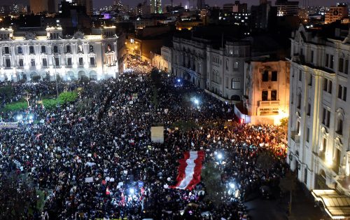 A large crowd of protestors fills a plaza and the surrounding streets at night. An enormous red and white flag floats over some of the participants.