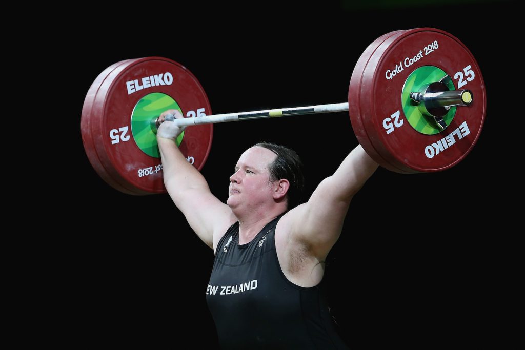 A woman in a black tank top with “New Zealand” written across the chest in white, holds a barbell with green and red weights on it.