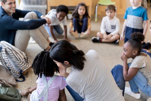 A group of young children and two adults sit in a circle. One of the adults whispers in a child’s ear while the others watch.