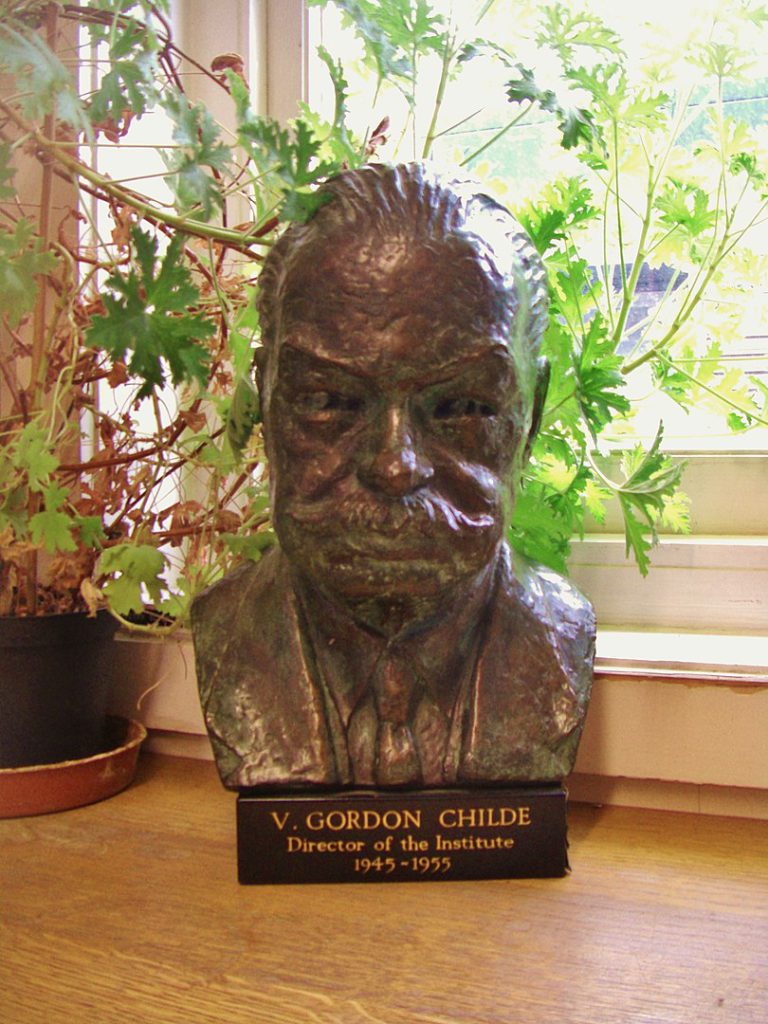 A bronze bust sits on a wood surface beside a plant and in front of a window pane.