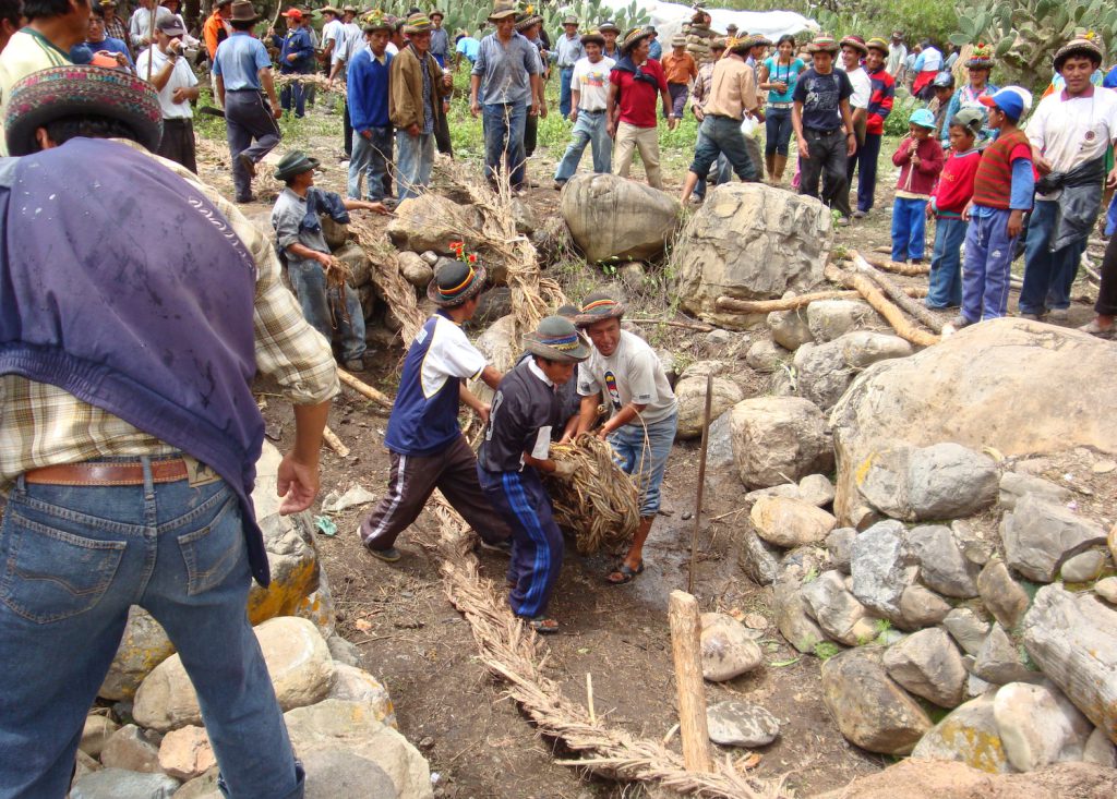 Community members look on as a group of people unfurl a long, large rope on the ground next to another rope. They are surrounded by large stones.