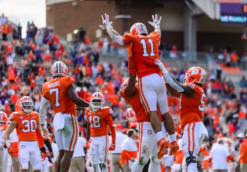 Two football players in orange and white uniforms lift their teammate in the air in celebration while surrounded by other team members.
